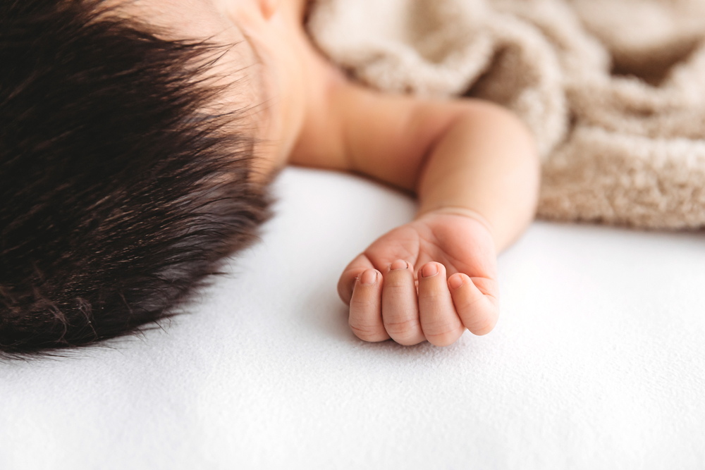 Sleeping newborn baby's tiny hand and dark hair on white blanket with cream knit wrap by professional photographer
