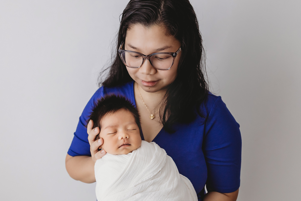 Adult woman in blue top holding sleeping newborn baby wrapped in white blanket, soft studio lighting by Tracy Rowell