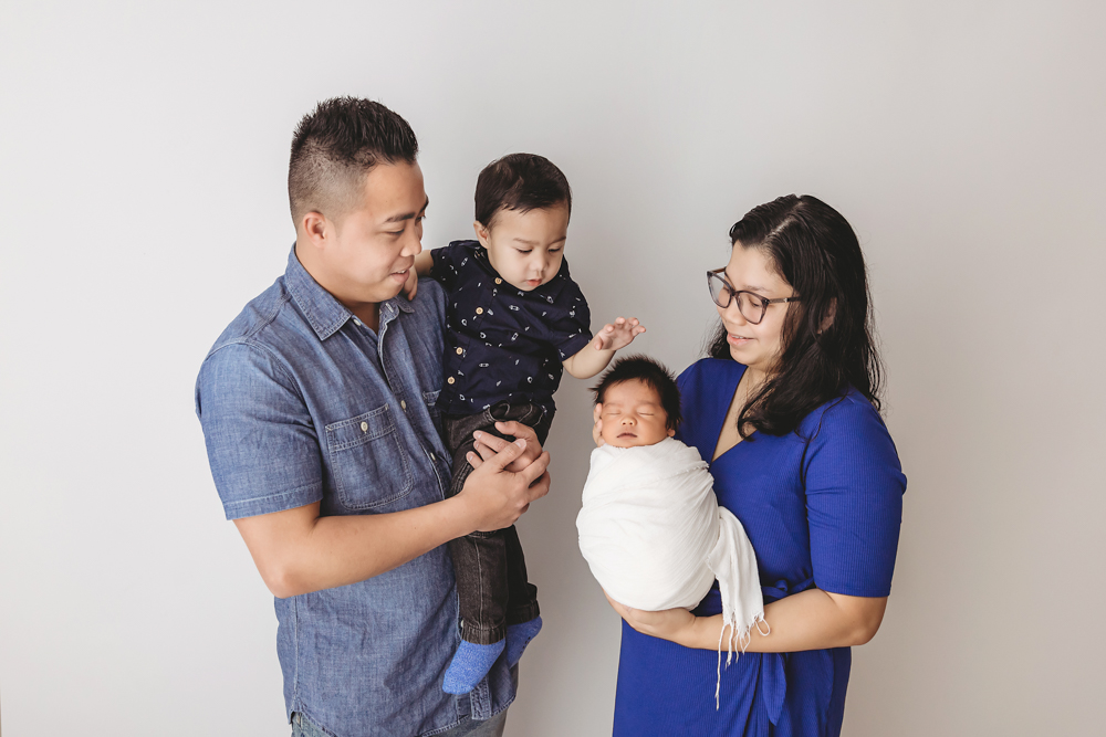 Family of four posing for newborn photos: dad in blue chambray shirt holding toddler, mom in royal blue dress cradling baby