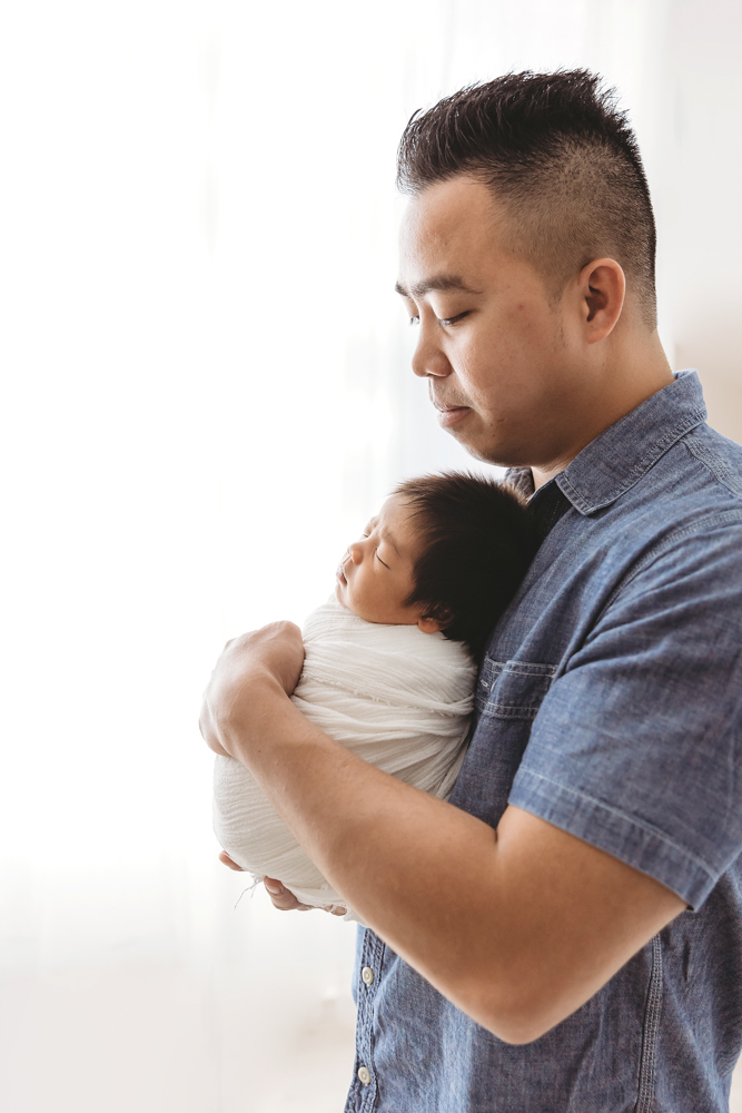 Father cradling sleeping baby in cream wrap, soft natural lighting, newborn pictures session in Florence SC