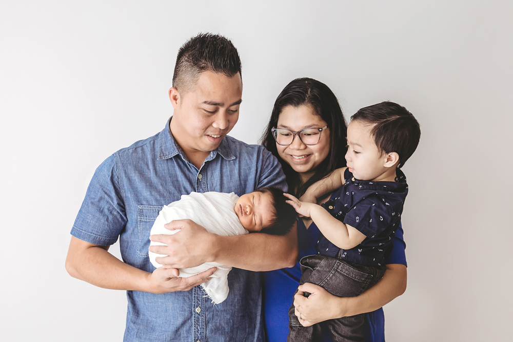 Family of four with newborn baby on light backdrop, parents in blue holding sleeping infant and toddler, florence sc photographer