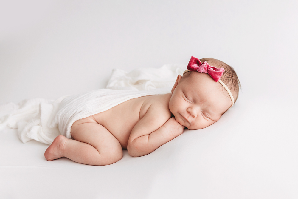 Newborn girl sleeps on ivory muslin wrap with a velvet rose headband by Reflection Images, Florence SC professional studio.