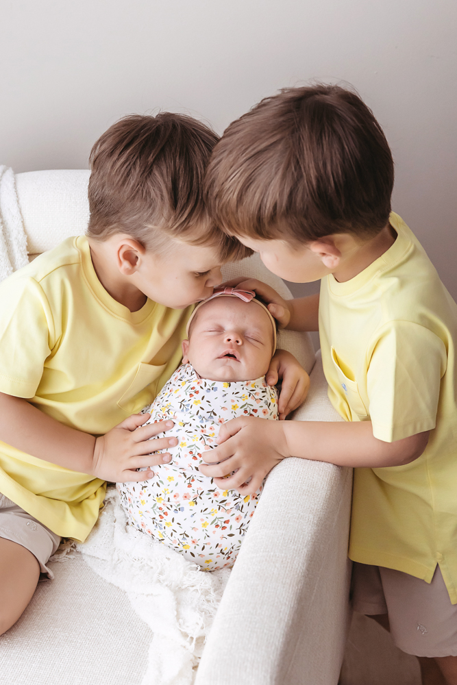 twin big brothers in yellow tees kiss newborn Gracyn, wrapped in a floral muslin swaddle, by Florence SC photographer Tracy...