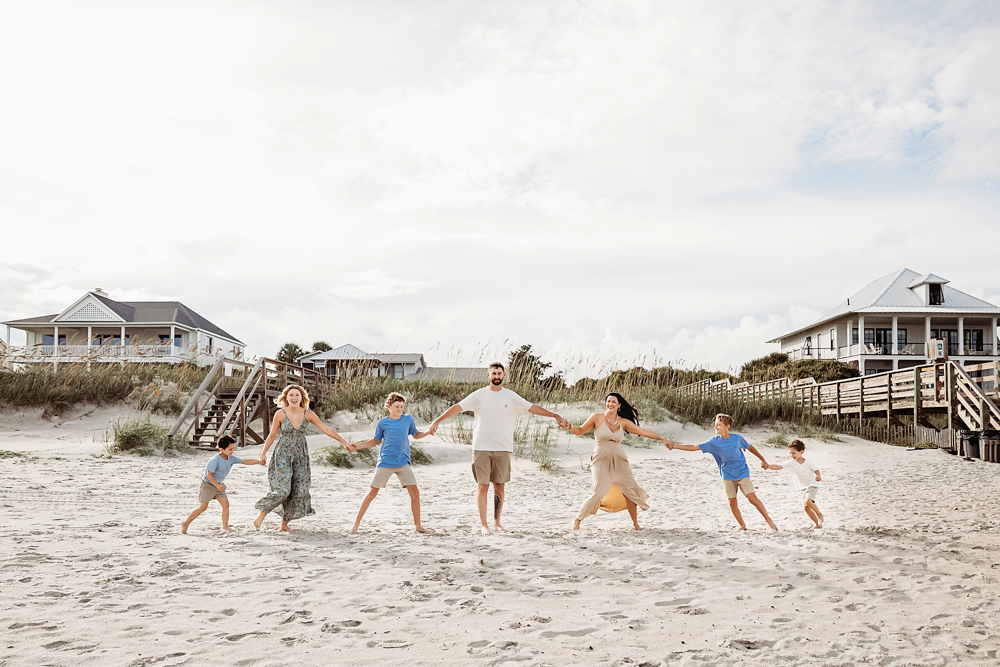 Family holding hands on Pawleys Island SC beach, wearing sage floral, azure blue, and tan linen as a beach photographer.