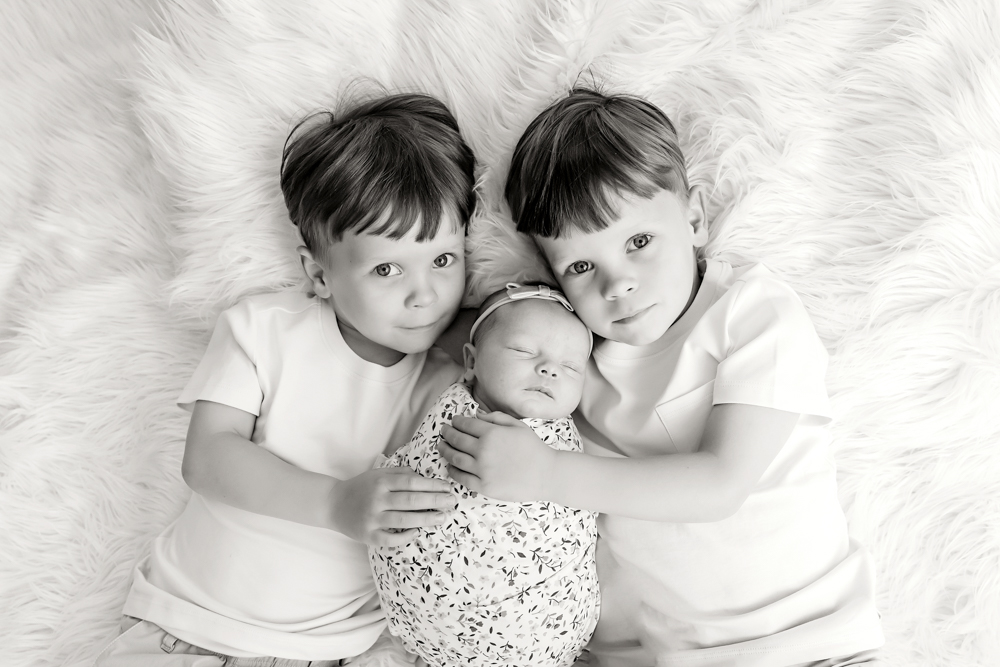 twin young brothers in white tees embrace a newborn in a floral swaddle on white faux fur by Florence SC Reflection Images sibling pose