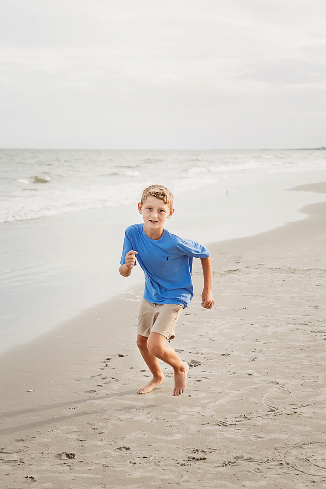 Boy in blue polo and khaki shorts running on the sand during a Pawleys Island SC beach family photography session.