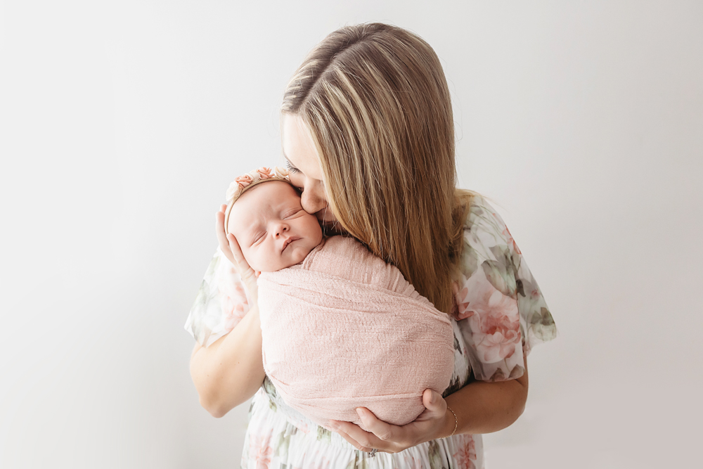Mother kissing sleeping newborn wrapped in dusty rose muslin, wearing floral headband, by Reflection Images Florence SC.
