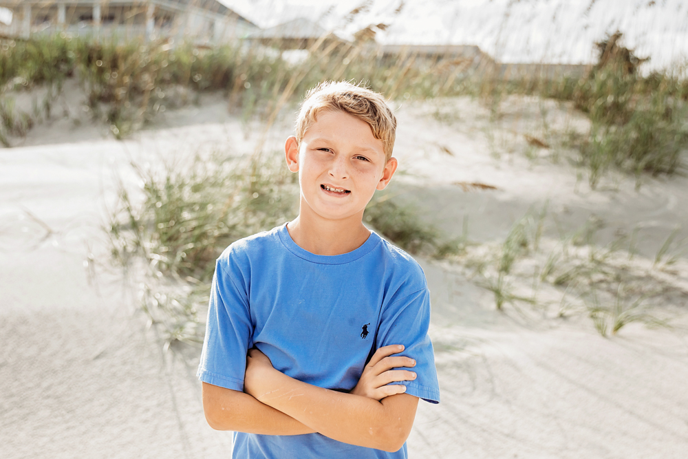 Boy in cornflower blue cotton t-shirt with arms crossed smiling at Pawleys Island SC beach family session.