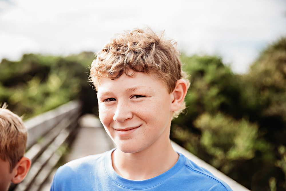 Boy with curly light brown hair and freckles smiling on a Pawleys Island SC boardwalk during family photography session.