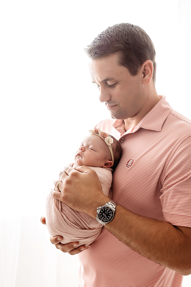 Father holds newborn girl in blush knit wrap with floral headband during Reflection Images by Tracy Rowell session,...