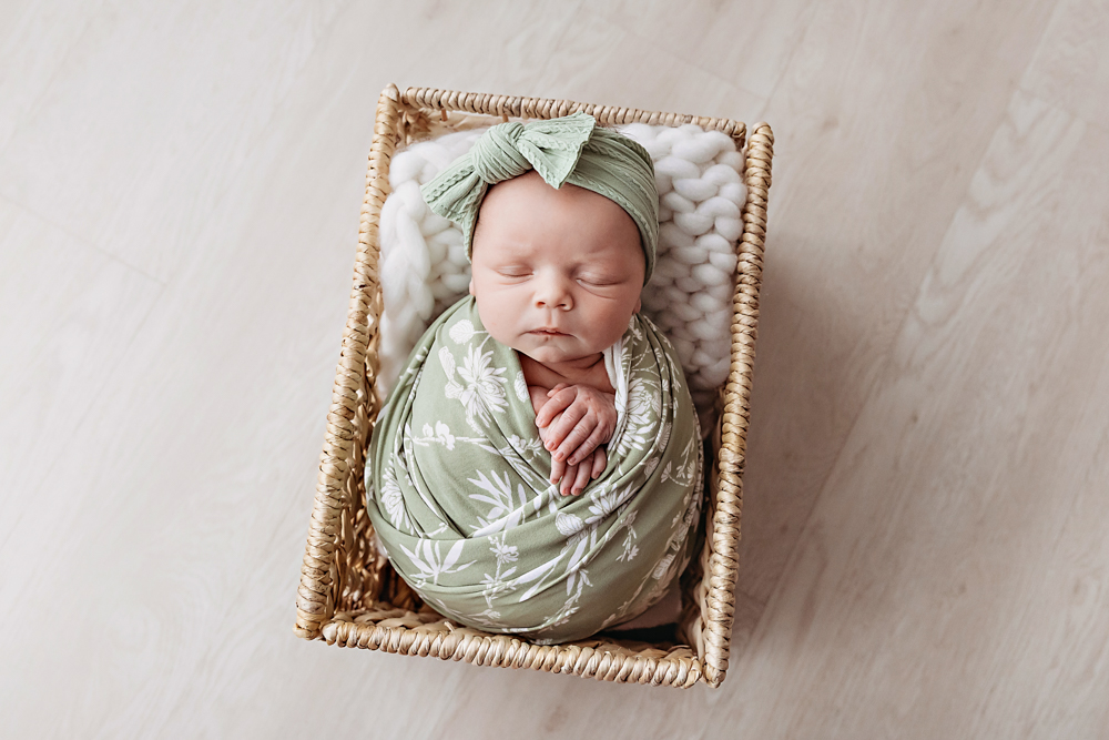 Gracyn in sage floral swaddle and headband inside a wicker basket on white chunky knit, by Reflection Images Florence SC.