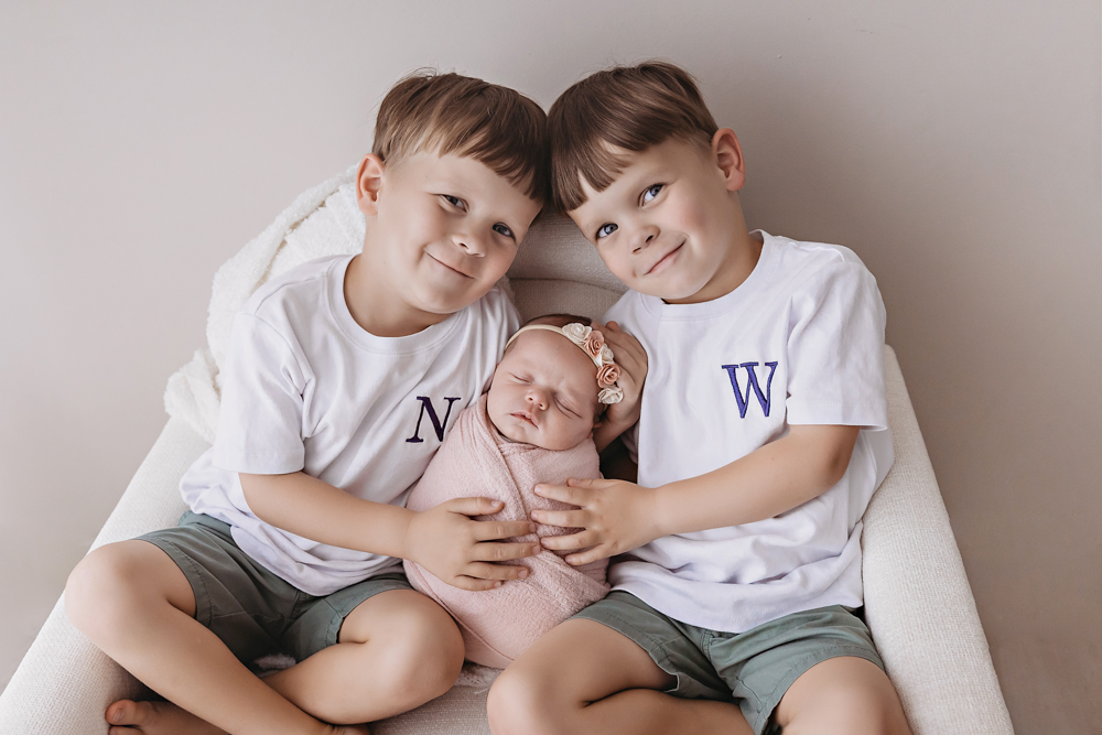 Two boys in white shirts and sage shorts holding a newborn in a dusty rose knit swaddle by Reflection Images Florence SC.
