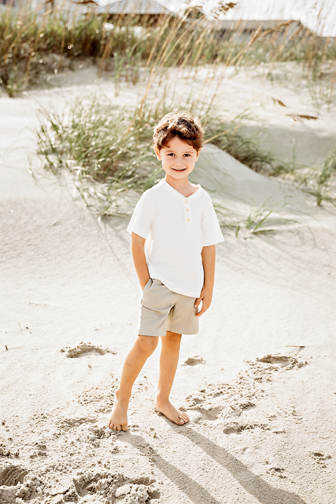 Smiling boy in white henley and taupe shorts posing on Pawleys Island SC sand dunes for beach family photography session.