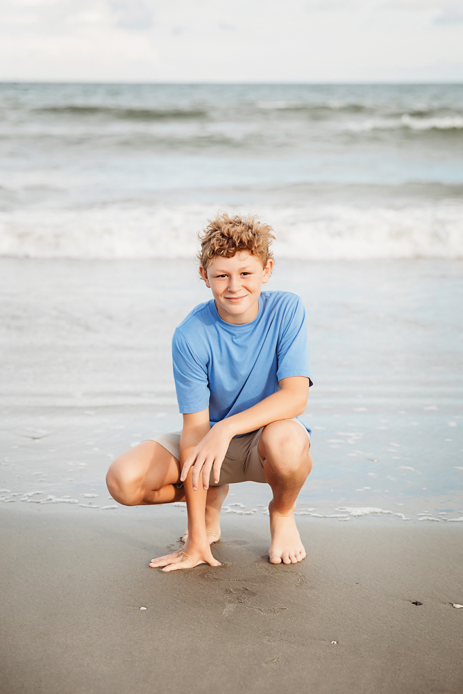 Smiling boy in cornflower blue t-shirt and khaki shorts crouching on Pawleys Island SC beach for family photography.