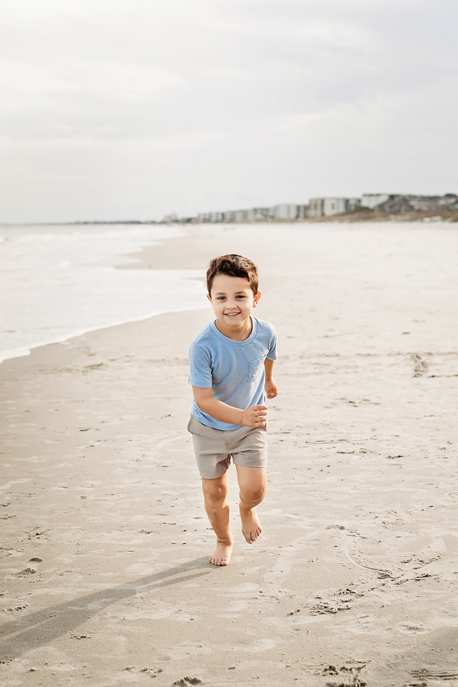 Smiling boy in powder blue t-shirt and taupe shorts running on Pawleys Island SC beach for family photography session.